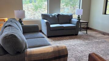 A living room with a grey sofa, a matching ottoman, and a small table with a lamp. There are two windows providing a view of a landscape.