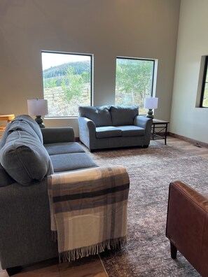 A living room with a grey sofa, a matching ottoman, and a small table with a lamp. There are two windows providing a view of a landscape. - Paradise Pines - Swap Screen Time With Family Time (Scofield)