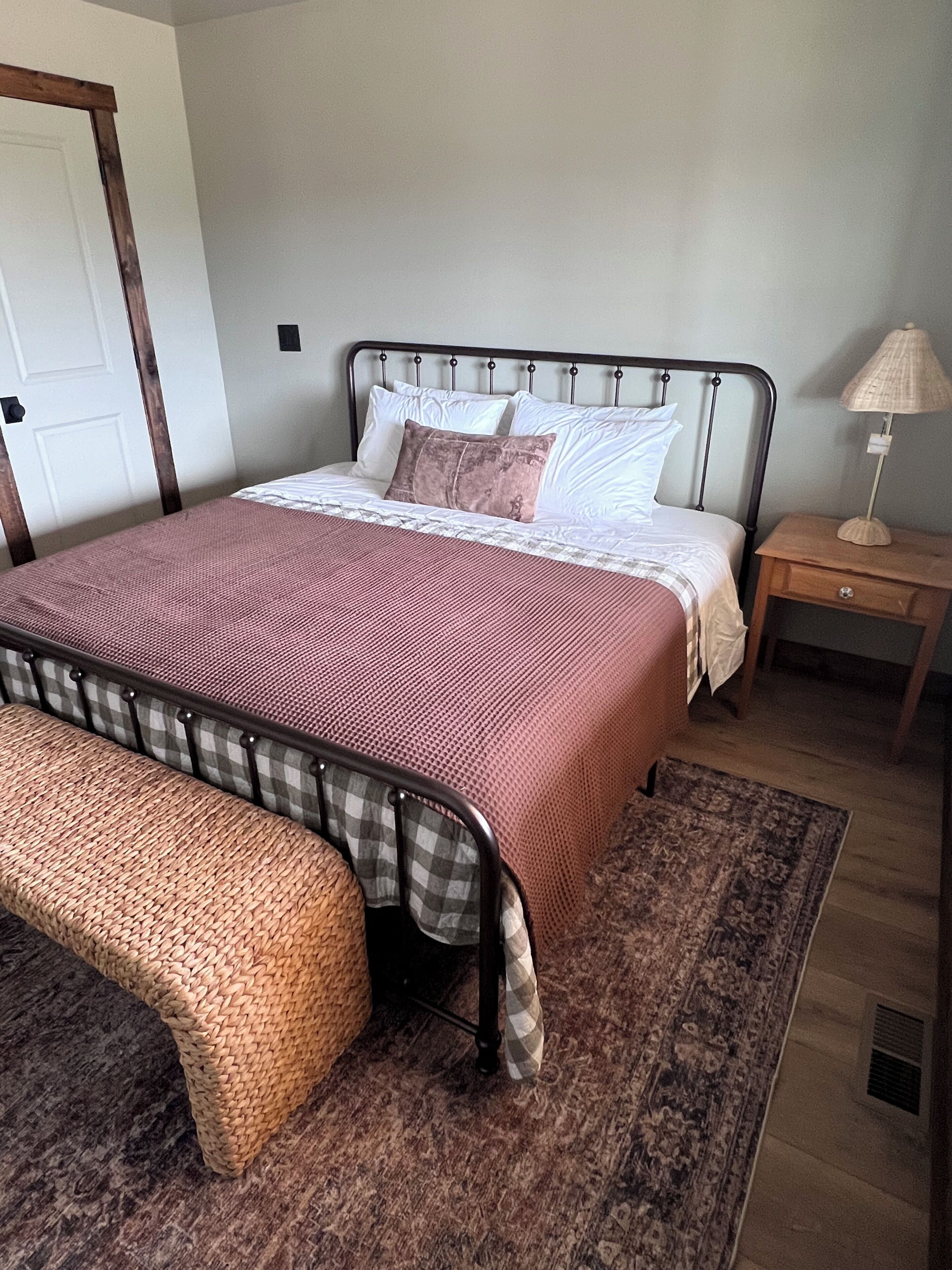 A bed with a metal frame, a wooden nightstand, a woven bench, and a patterned rug.