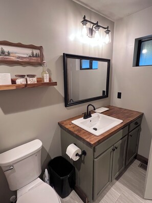 A bathroom with a white toilet, a sink with a wooden countertop, a mirror, and a black trash bin. - Paradise Pines - Swap Screen Time With Family Time (Scofield)