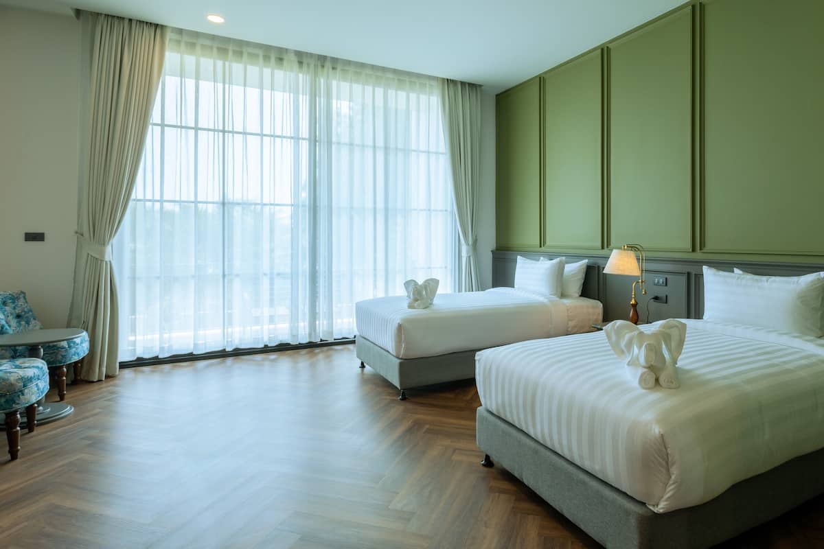 A hotel room with two beds, a table, and a window with sheer curtains.