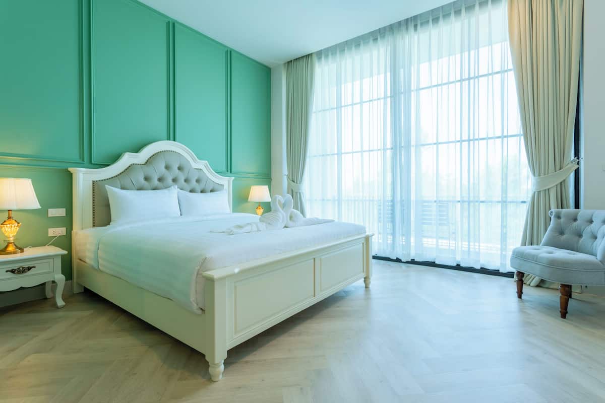 A large bed with a tufted headboard, two bedside tables with lamps, a chair, and a large window with sheer curtains.
