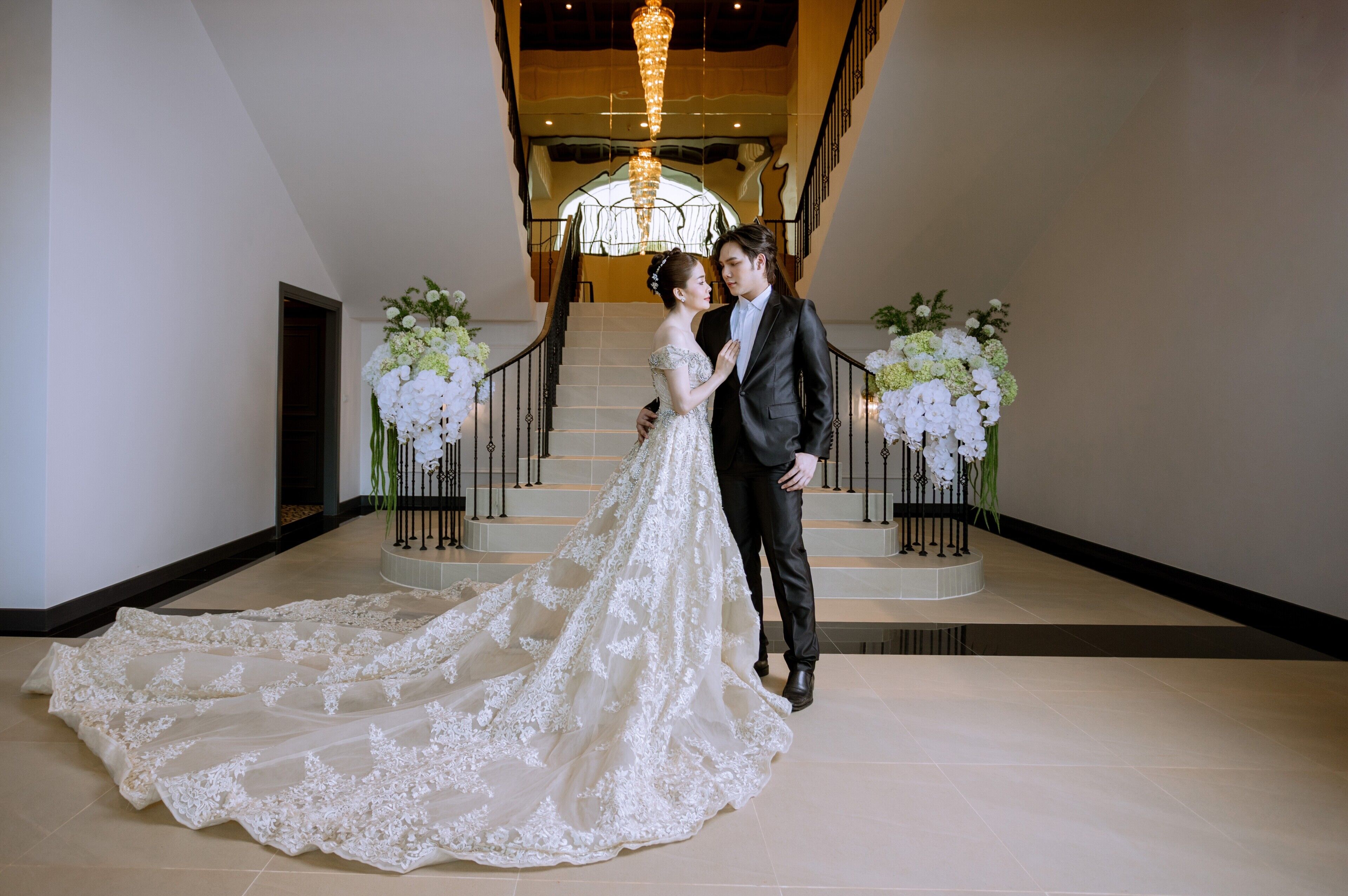 A couple in wedding attire standing in a grand staircase area with floral arrangements and a chandelier.