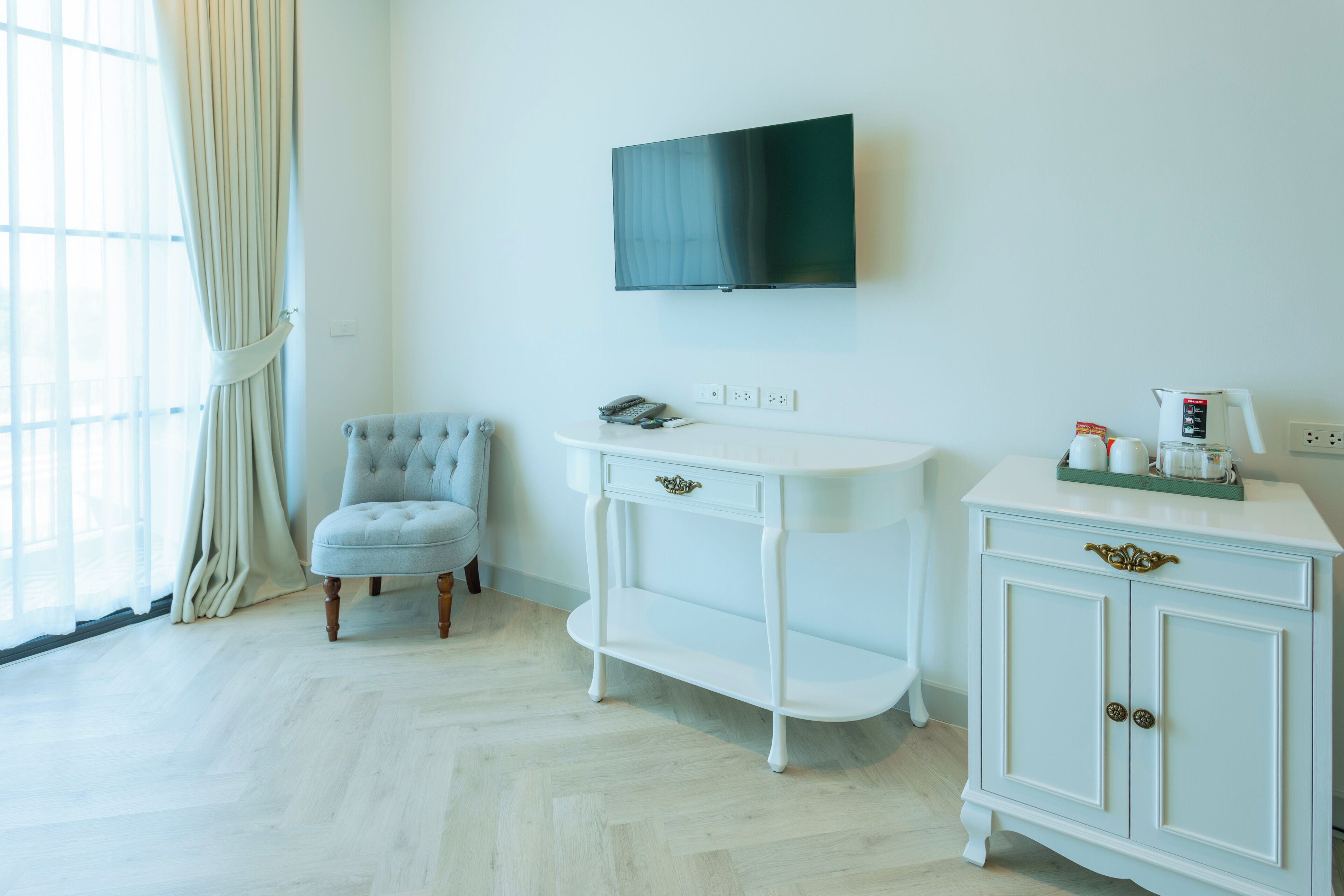 Una habitación con un tocador blanco, un sillón capitonado, un televisor montado en la pared y una ventana con cortinas.