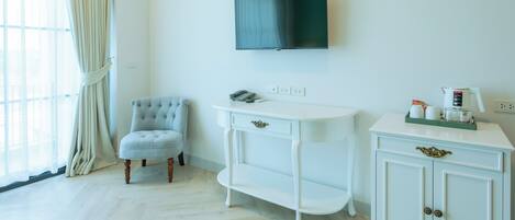 Una habitación con un tocador blanco, un sillón capitonado, un televisor montado en la pared y una ventana con cortinas.