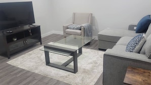 Un salon moderne avec un téléviseur à écran plat, une table basse en verre, un canapé gris agrémenté de coussins à motifs, un fauteuil beige et un meuble de rangement en bois foncé pour les appareils électroniques.