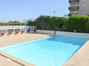 Una piscina rectangular con una escalera metálica, rodeada por un deck con baldosas color beige. A lo largo del borde de la piscina hay macetas con flores rosas. Se observa un seto y un edificio con balcones al fondo.