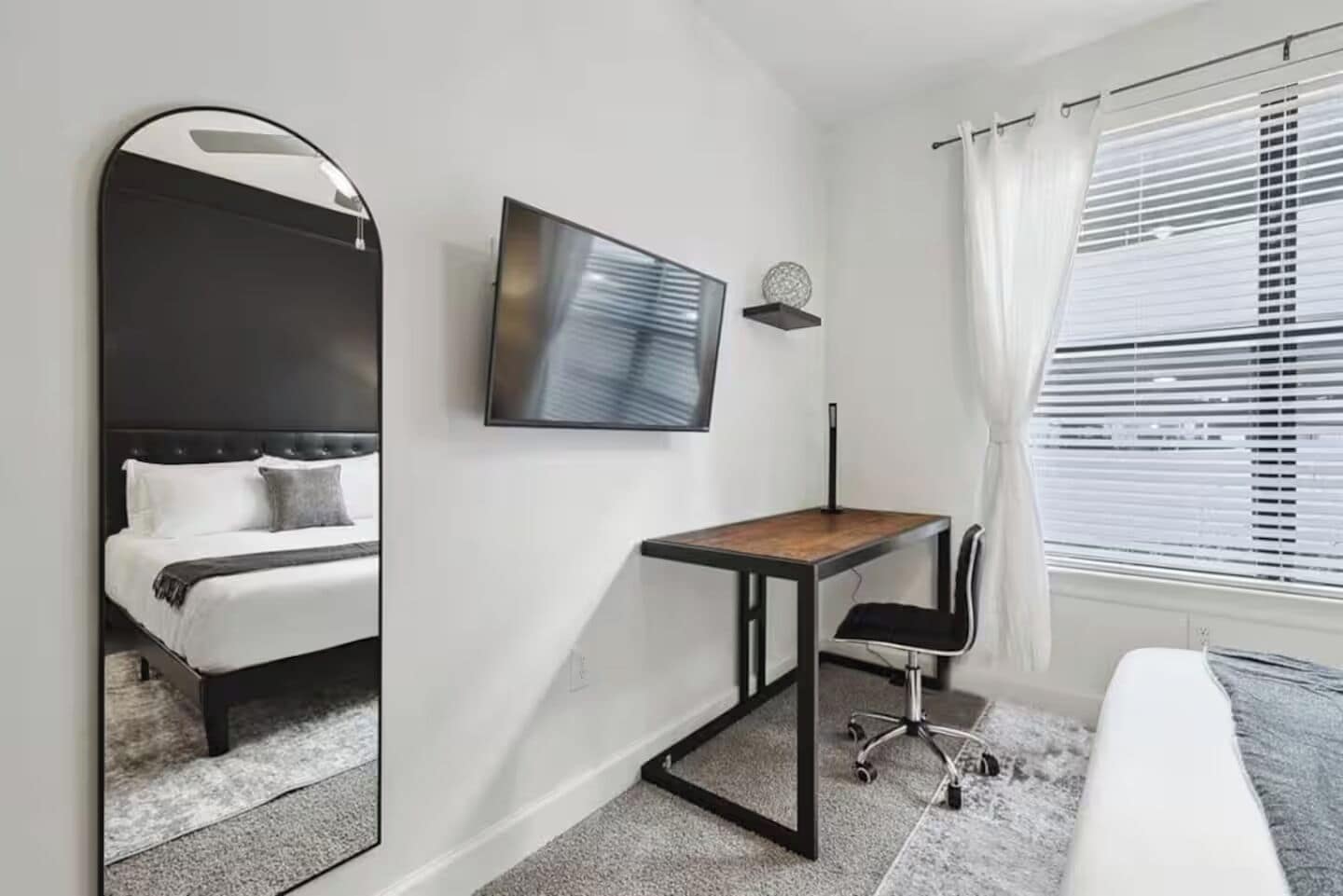 Moderni hotellihuone, jossa on sänky, työpöytä, tuoli, suuri peili, taulutelevisio ja ikkuna, jossa on sälekaihtimet.