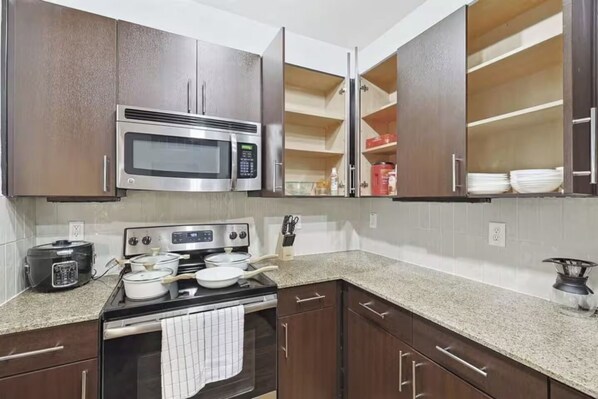 A kitchen with dark wooden cabinets, a stainless steel microwave, a stove, and a countertop with a rice cooker and pots. - Chic and Stylish 2B near Legacy West  (Frisco)