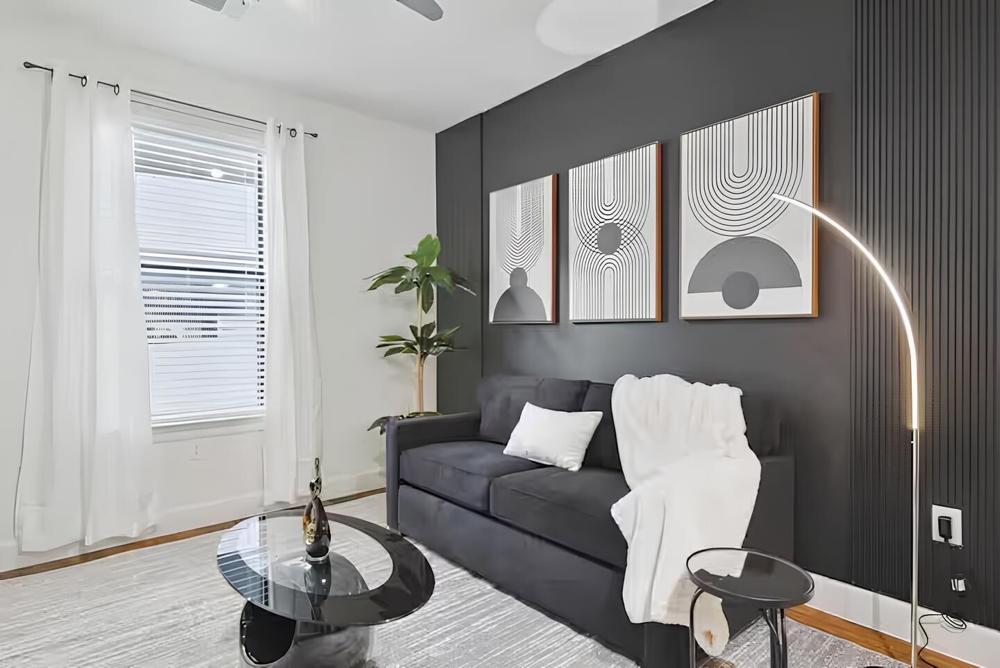Un salon moderne avec un canapé gris foncé, une table basse en verre et un lampadaire. Des œuvres d’art abstraites ornent le mur au-dessus du canapé, et une plante en pot se trouve à gauche.