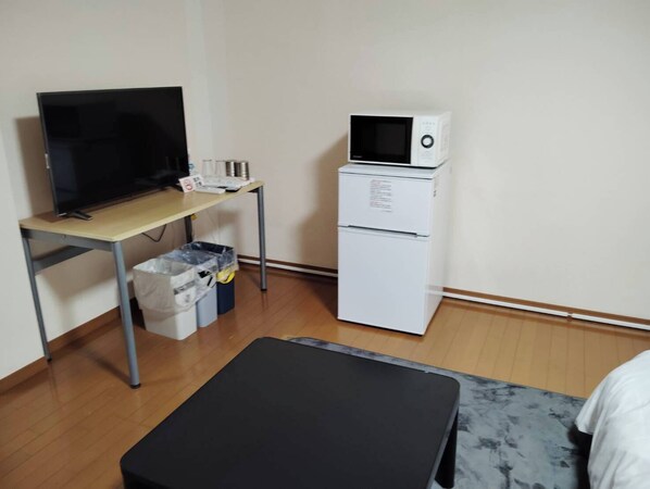 A small room with a desk, a television, a microwave, and a trash bin.