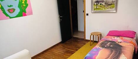 A room with a white sofa, a bed with colorful bedding, a small stool, and a door leading to another room.