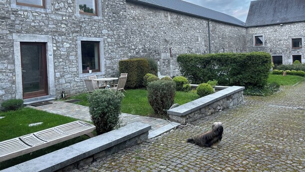 Un edificio de piedra con un patio empedrado, una mesa pequeña y sillas, y un perro en el camino adoquinado.