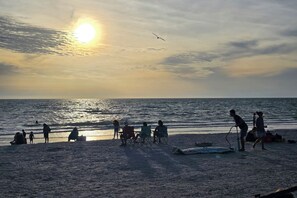A beach scene with people sitting and standing by the water, a clear sky, and the sun setting or rising. - Southern PAG Beach House Bungalows (St. Pete Beach)