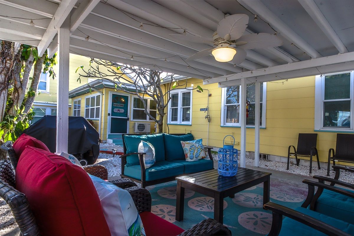 An outdoor patio area with a covered seating arrangement, a grill, and a yellow building in the background.
