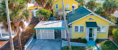 A yellow house with a blue roof, surrounded by palm trees and a white fence.