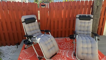 Two lounge chairs with footrests on a patterned outdoor rug, set against a wooden fence.