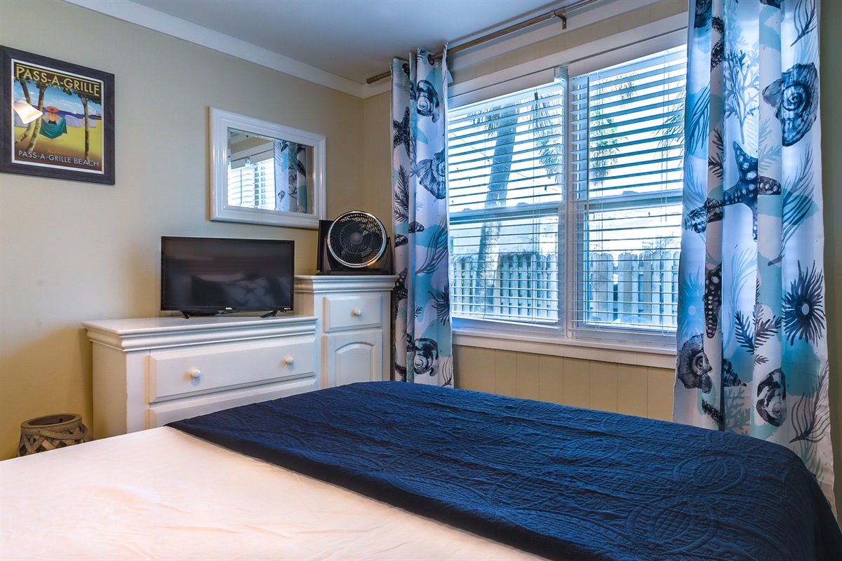 Un dormitorio con cama, televisor sobre una cómoda, ventana con persianas y un cartel con motivos playeros en la pared.
