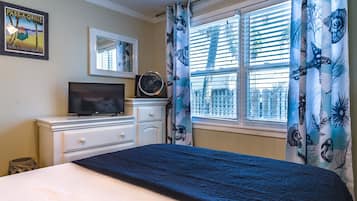 Un dormitorio con cama, televisor sobre una cómoda, ventana con persianas y un cartel con motivos playeros en la pared.