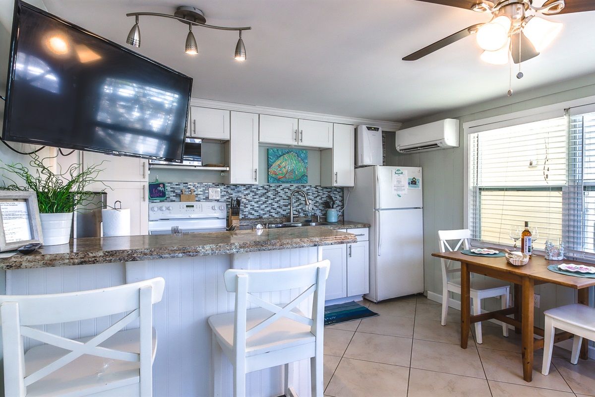 A kitchen with a flat-screen TV, white cabinets, a granite countertop, and a dining area with a wooden table and white chairs.