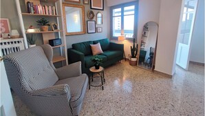 Ein Wohnzimmer mit einem grünen Sofa, einem Hundezahn-Sessel, einem Bücherregal, einem Spiegel und einem Fenster mit Tageslicht.