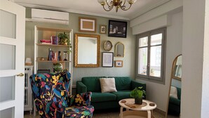 Ein Wohnzimmer mit einem grünen Sofa, einem farbenfrohen Sessel, einem kleinen runden Tisch und einem Bücherregal.