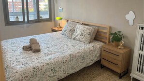 Ein Schlafzimmer mit einem Bett, einem Nachttisch, einer Lampe, einem Fenster und Blick auf Gebäude.