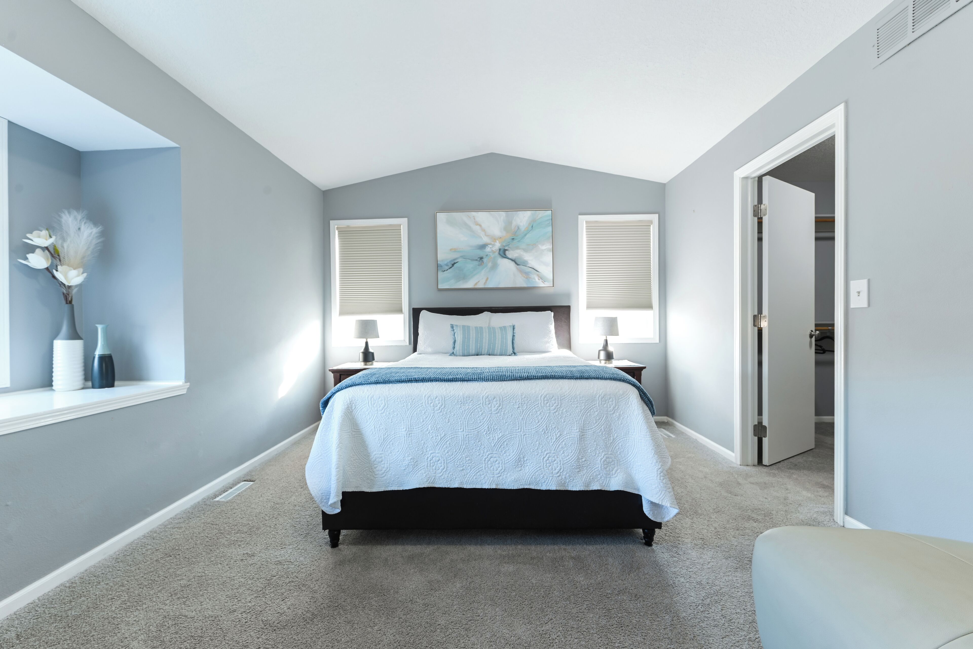 A bedroom with a bed centered in the room, flanked by two bedside tables with lamps, a window with blinds, and a door leading to another room.