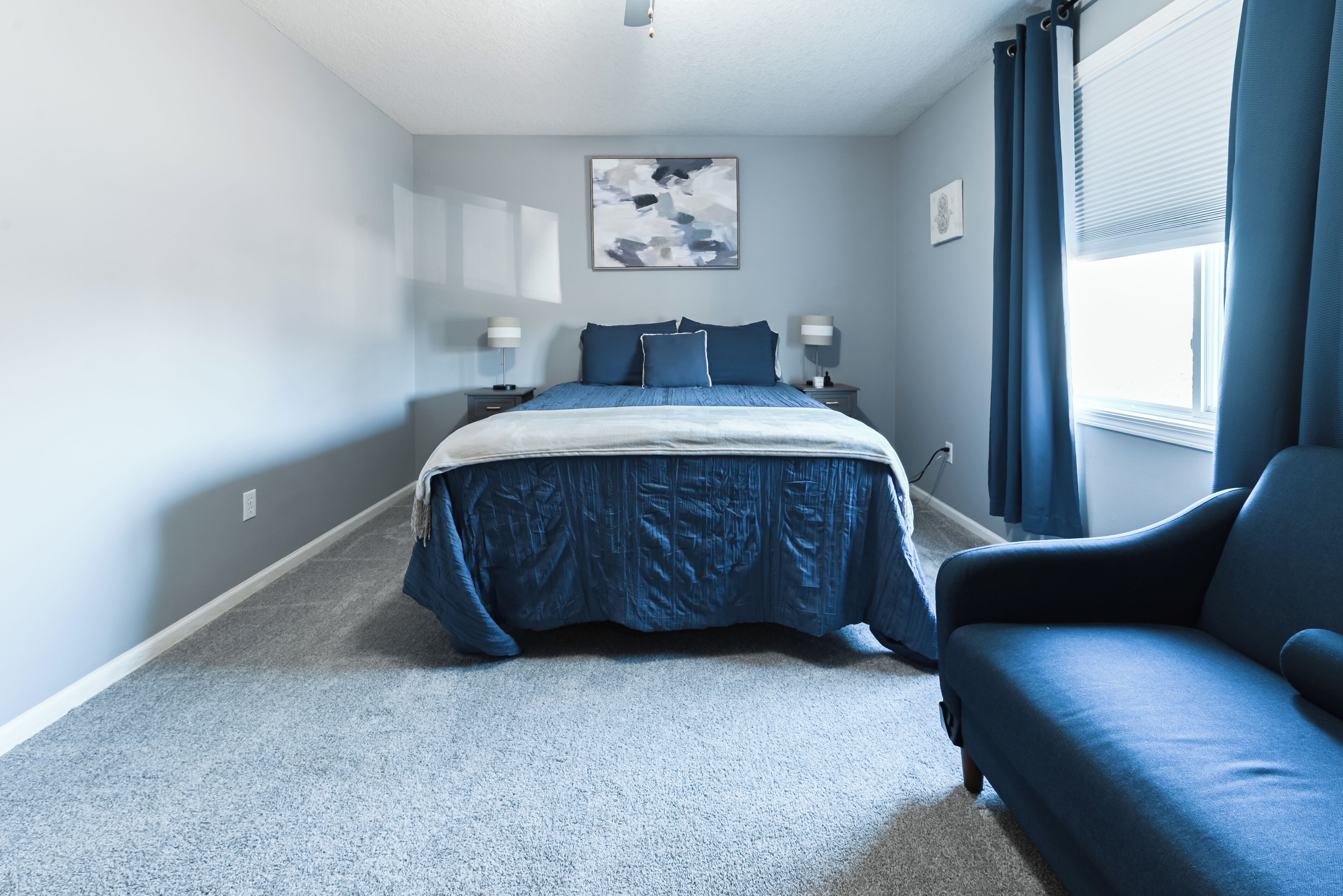Quarto com cama, duas mesas de cabeceira, janela com cortinas e um sofá azul.