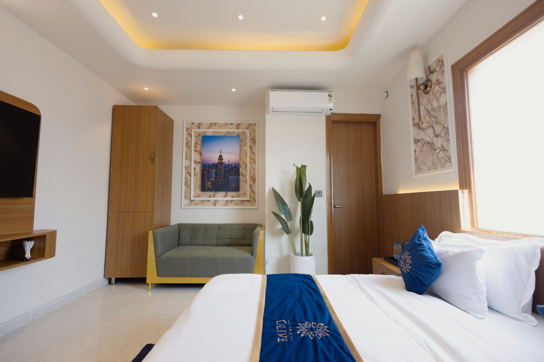 A modern hotel room with a bed, a sofa, a potted plant, and a cityscape picture on the wall.