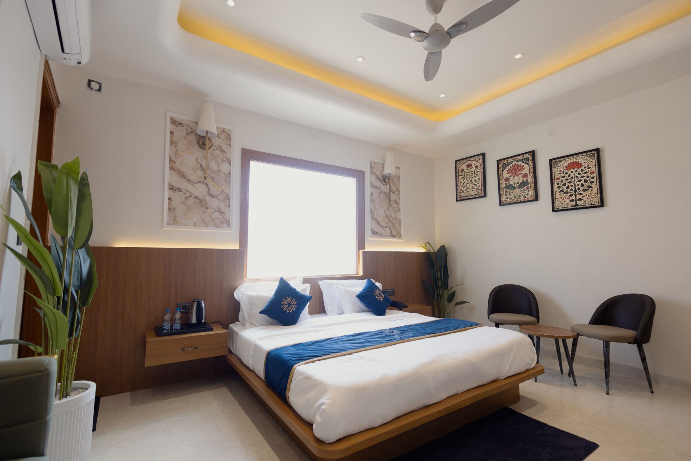 Une chambre d’hôtel moderne dotée d’un grand lit, de tables de chevet, d’un bureau, de deux chaises, d’un ventilateur de plafond et d’œuvres d’art accrochées aux murs.
