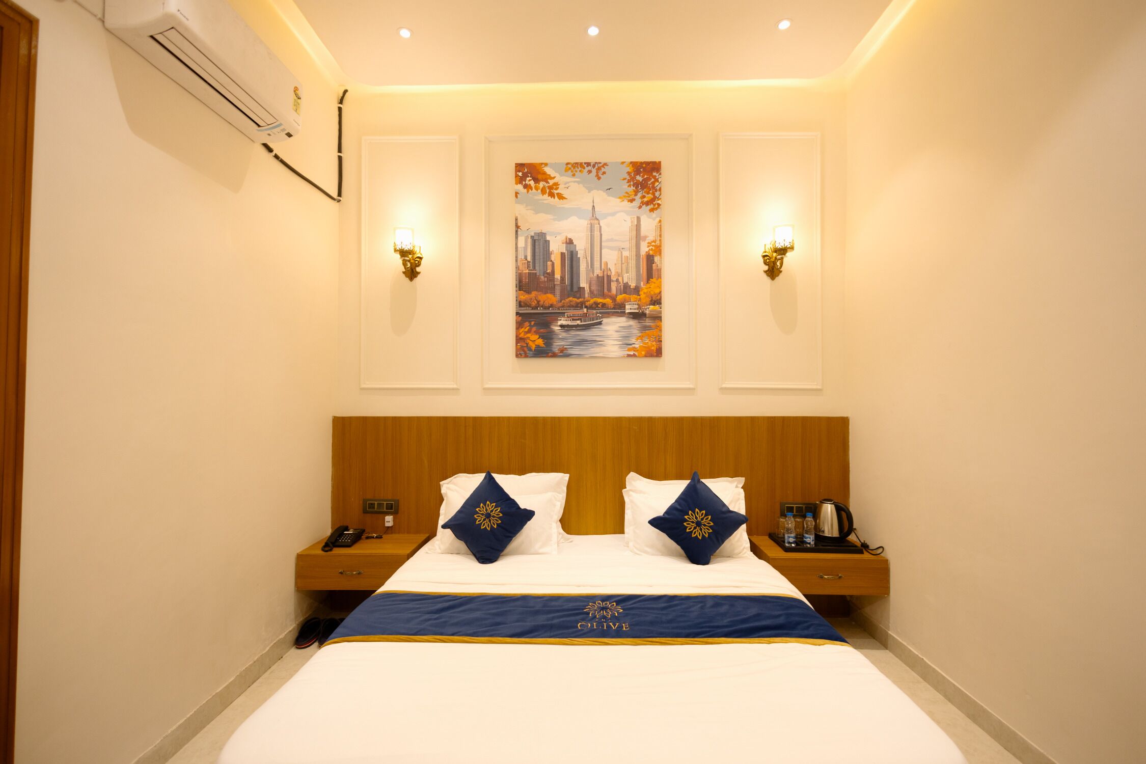 A hotel room with a bed, two bedside tables, a nightstand, an air conditioning unit, and a painting of a cityscape.