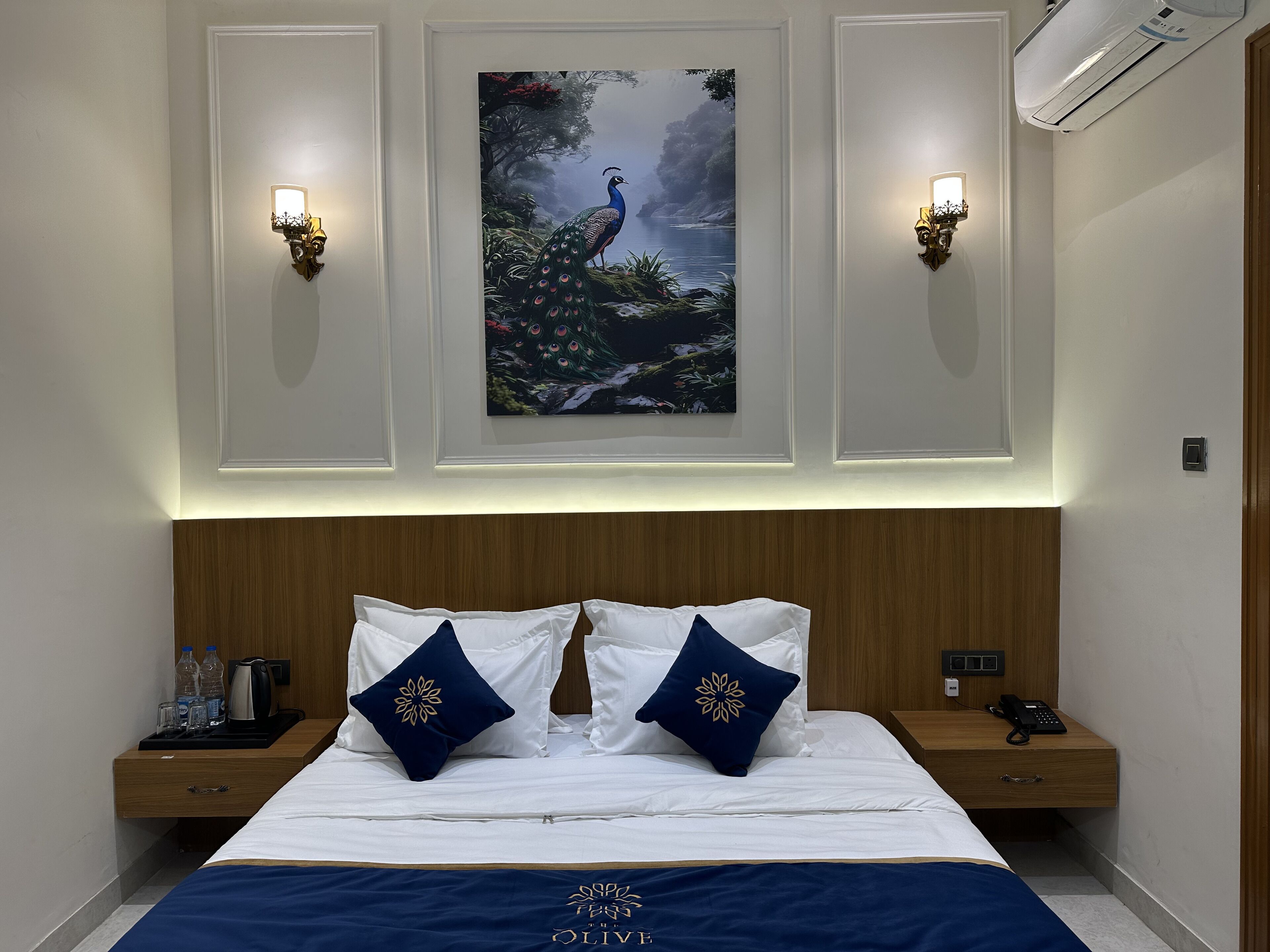 Ein Hotelzimmer mit einem ordentlich bezogenen Bett, zwei Nachttischen mit Telefon und Wasserkocher, sowie einem Pfauengemälde an der Wand.