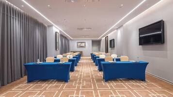 A conference room with rows of tables and chairs, a flat-screen TV on the wall, and a patterned carpet.