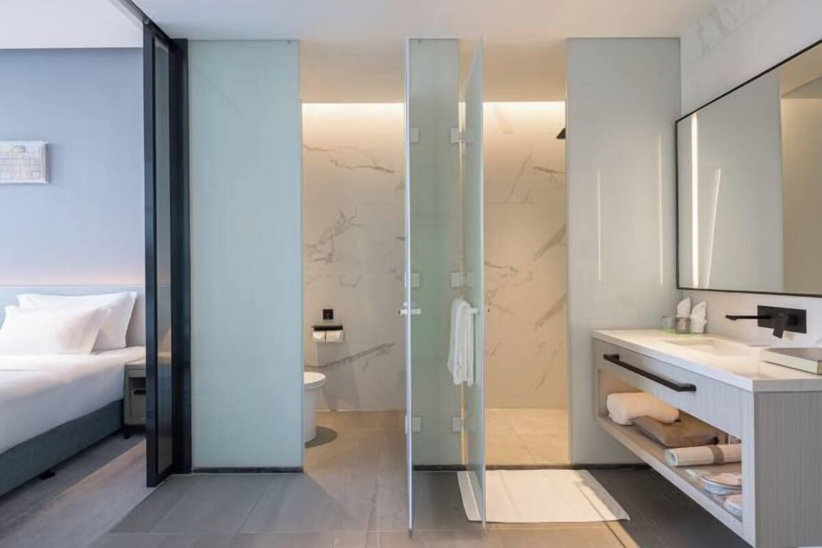 A modern hotel room with a glass shower enclosure, a bed, a bathroom with a marble wall, and a large mirror.