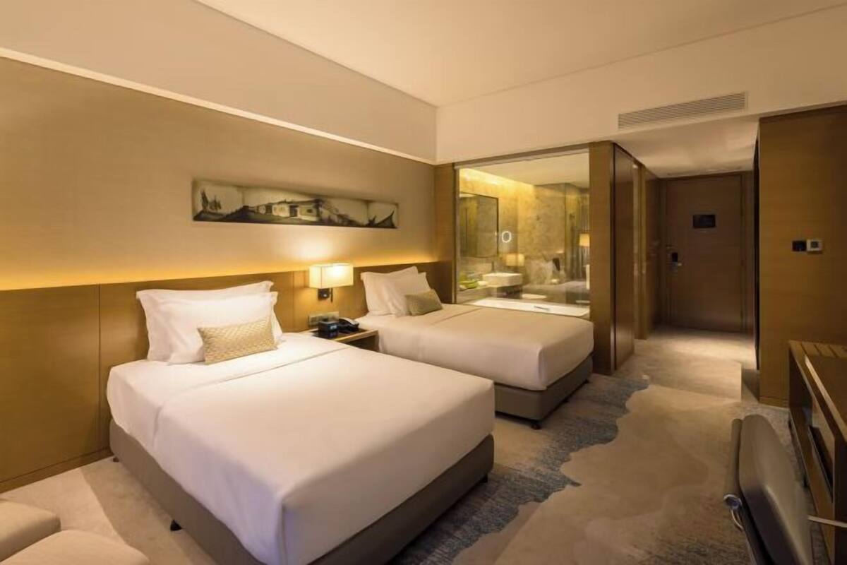 A hotel room with two beds, a desk, a chair, a painting on the wall, and a bathroom visible through a glass door.