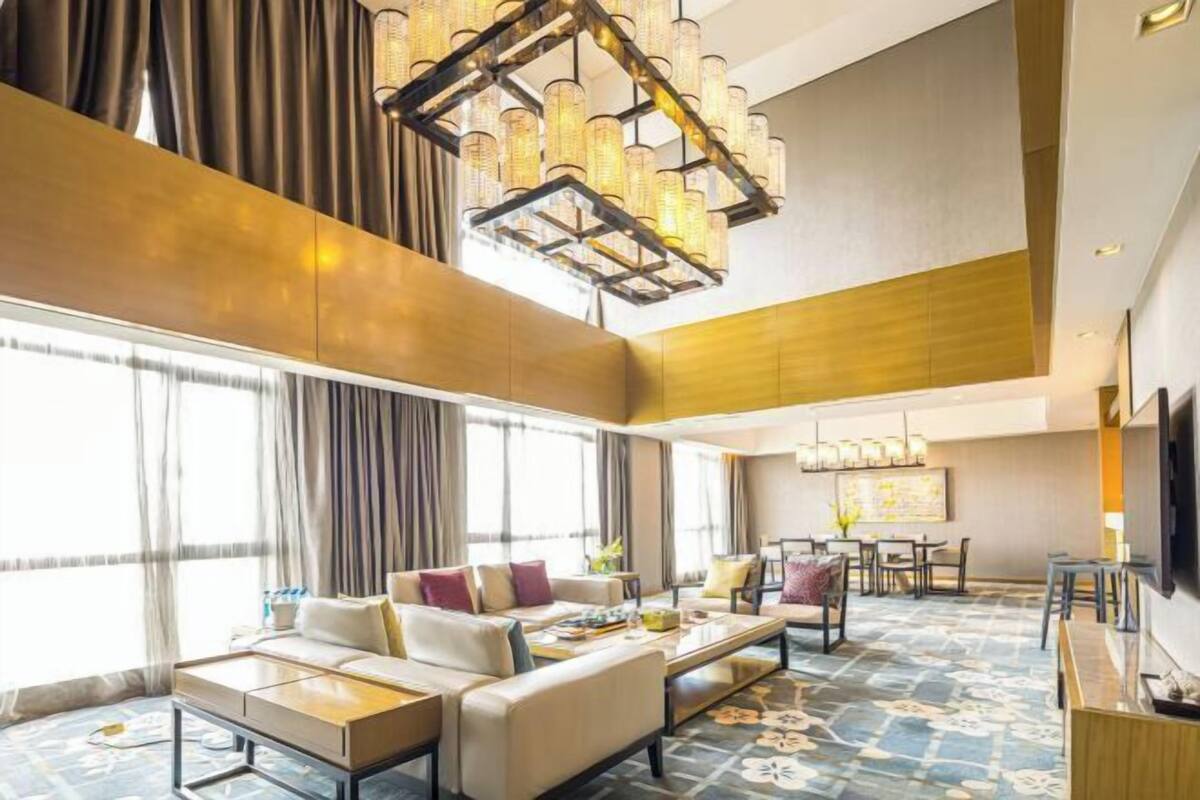 A spacious hotel lobby with a high ceiling, a large chandelier, a seating area with sofas and a coffee table, and a dining area with chairs and a table.