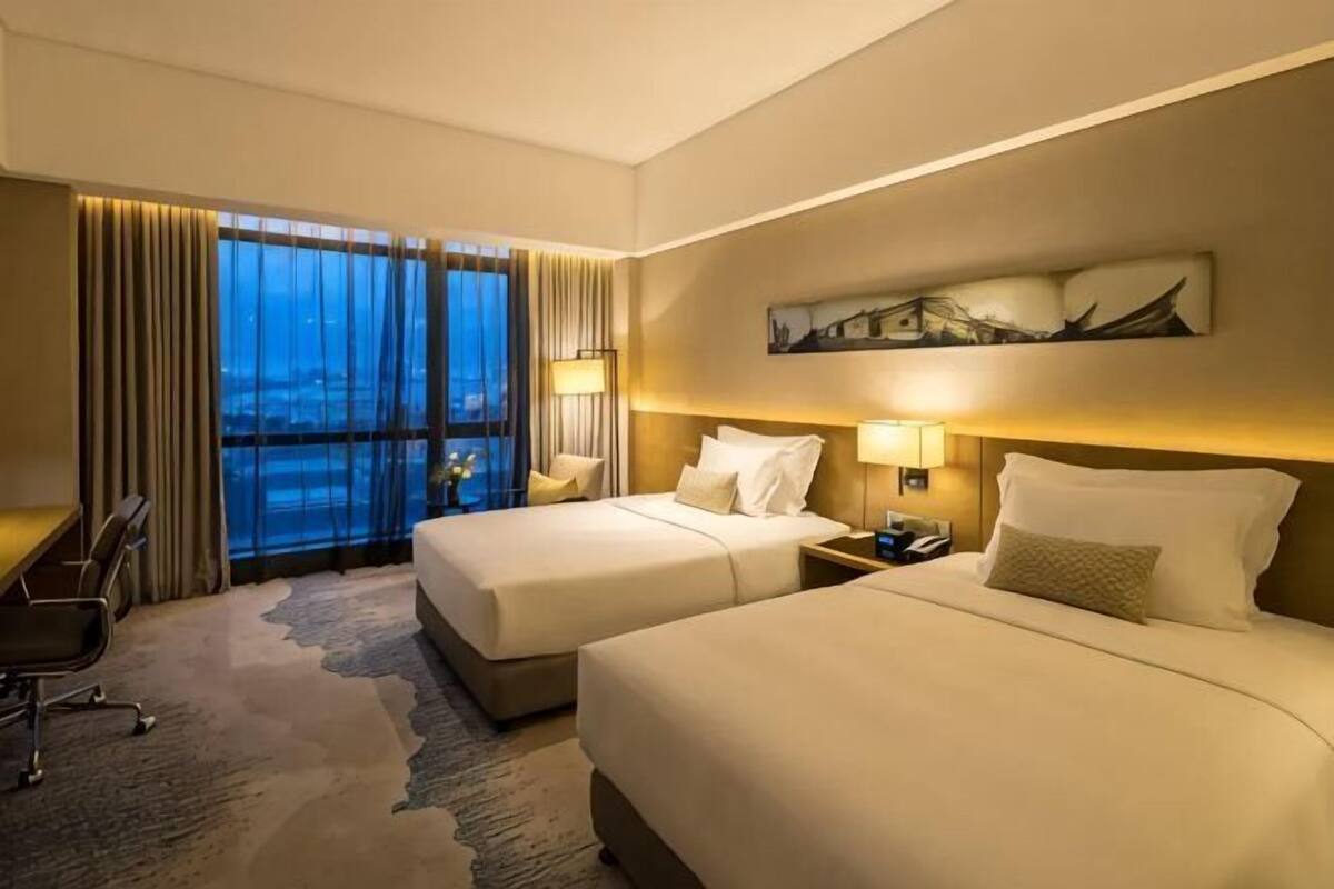 A hotel room with two beds, a desk, and a view of the city at dusk.