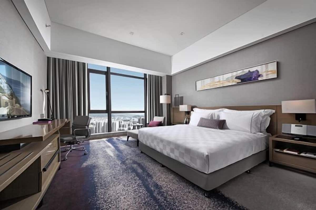 A modern hotel room with a large bed, a desk, a chair, a TV, and a city view.
