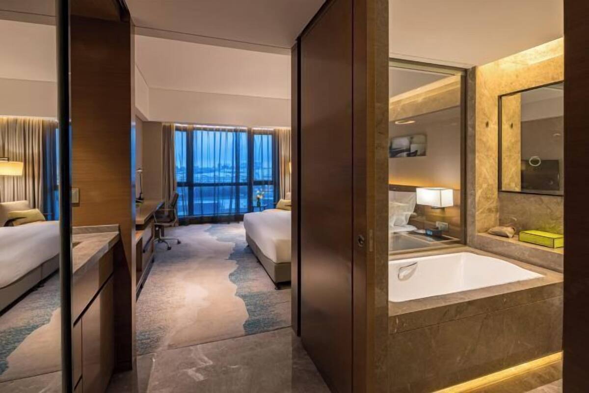 A hotel room with a large bed, a sitting area, a bathroom with a bathtub, and a view of the city.
