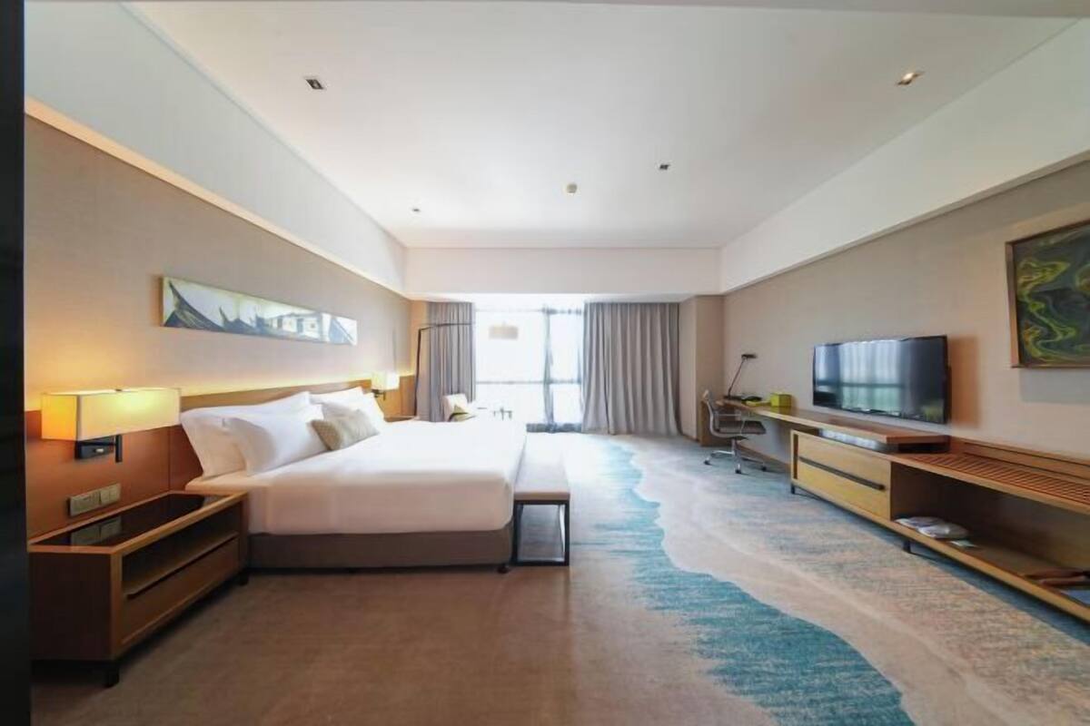 A hotel room with a bed, a TV, a desk, and a large window.