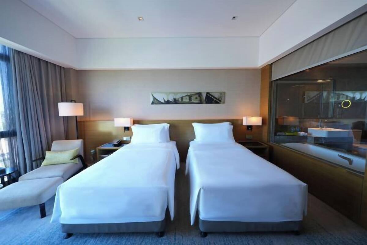 A hotel room with two single beds, a sitting area with a chair, a desk, and a large window with curtains.