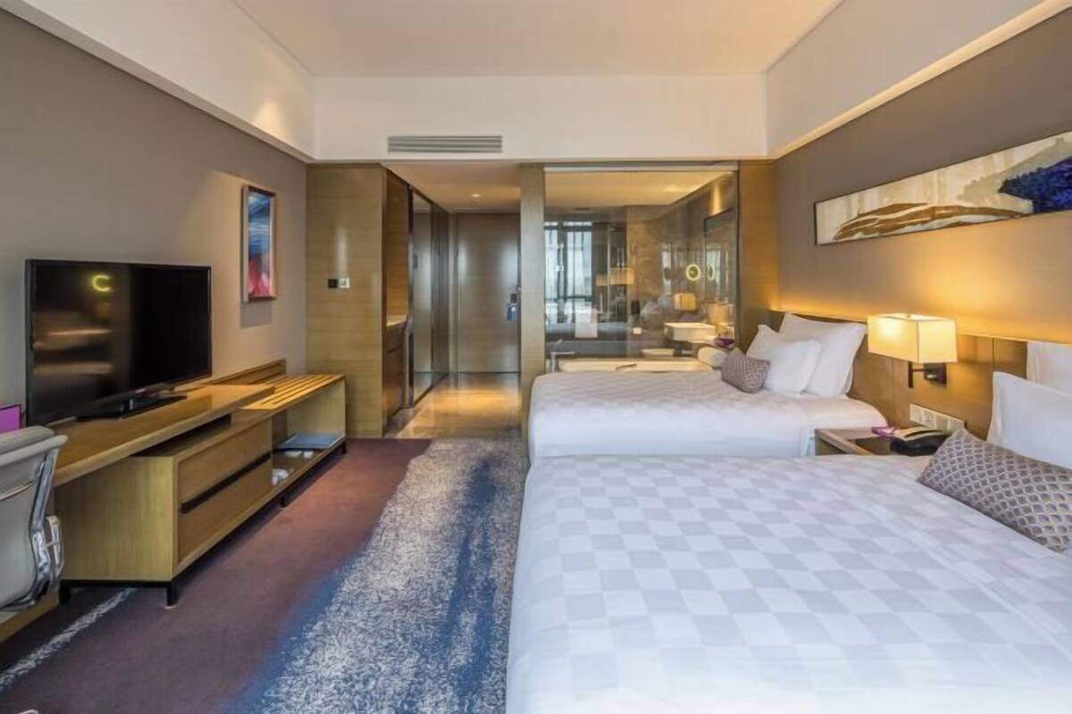 A hotel room with two beds, a TV, a desk, and a bathroom visible through a glass door.
