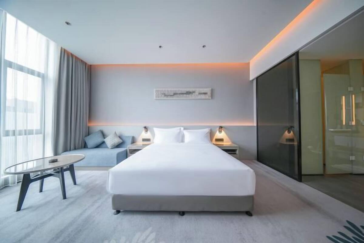 A modern hotel room with a large bed, bedside tables with lamps, a small round table, and a large window with curtains.