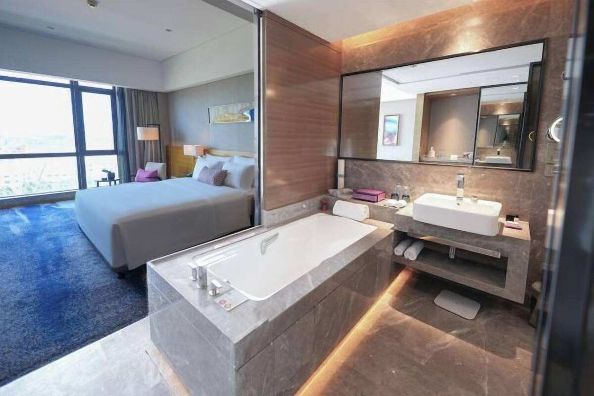 A modern hotel room with a large bed, a separate bathroom with a bathtub and two sinks, and a large window offering a city view.