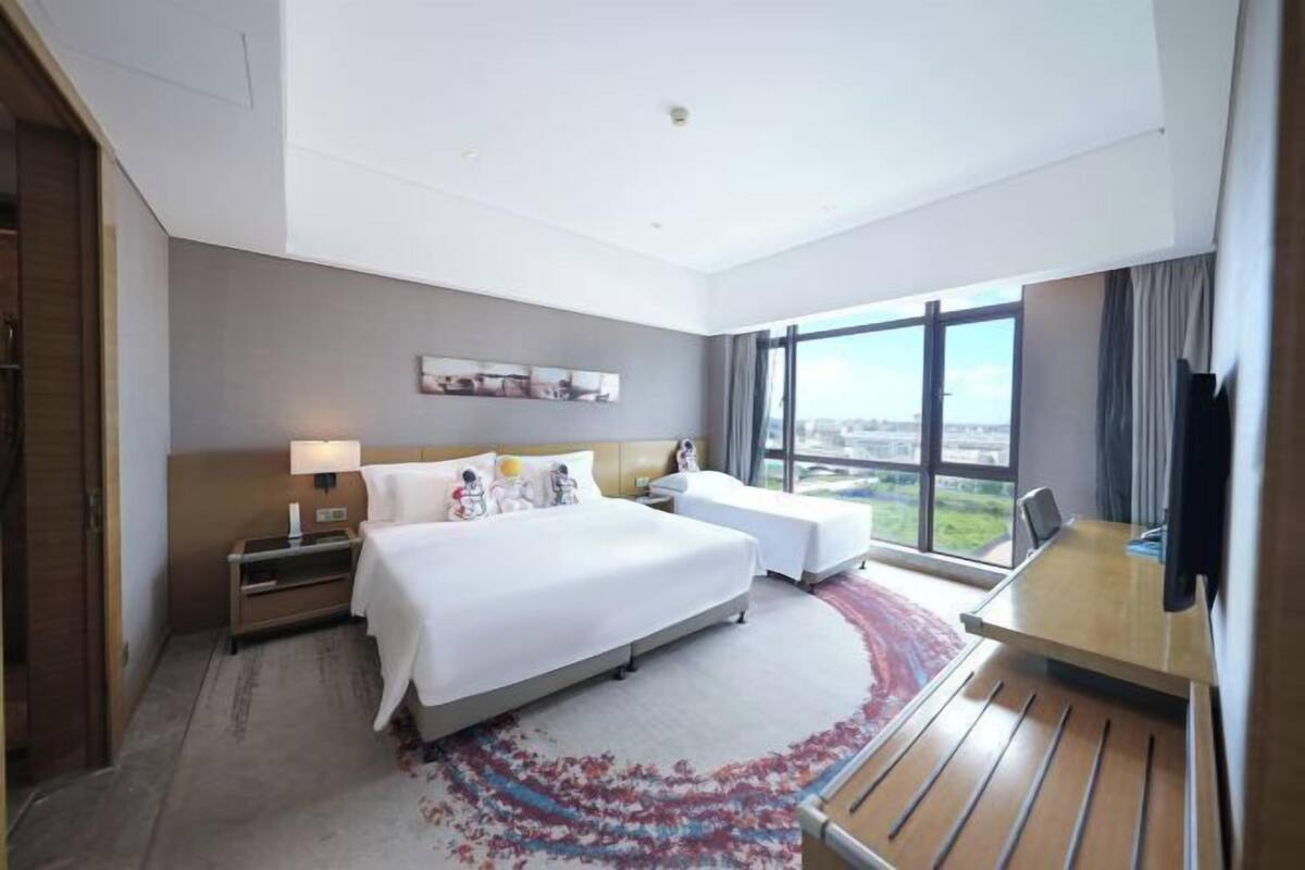 A hotel room with a large bed, a desk, a chair, a nightstand, and a window with a city view.