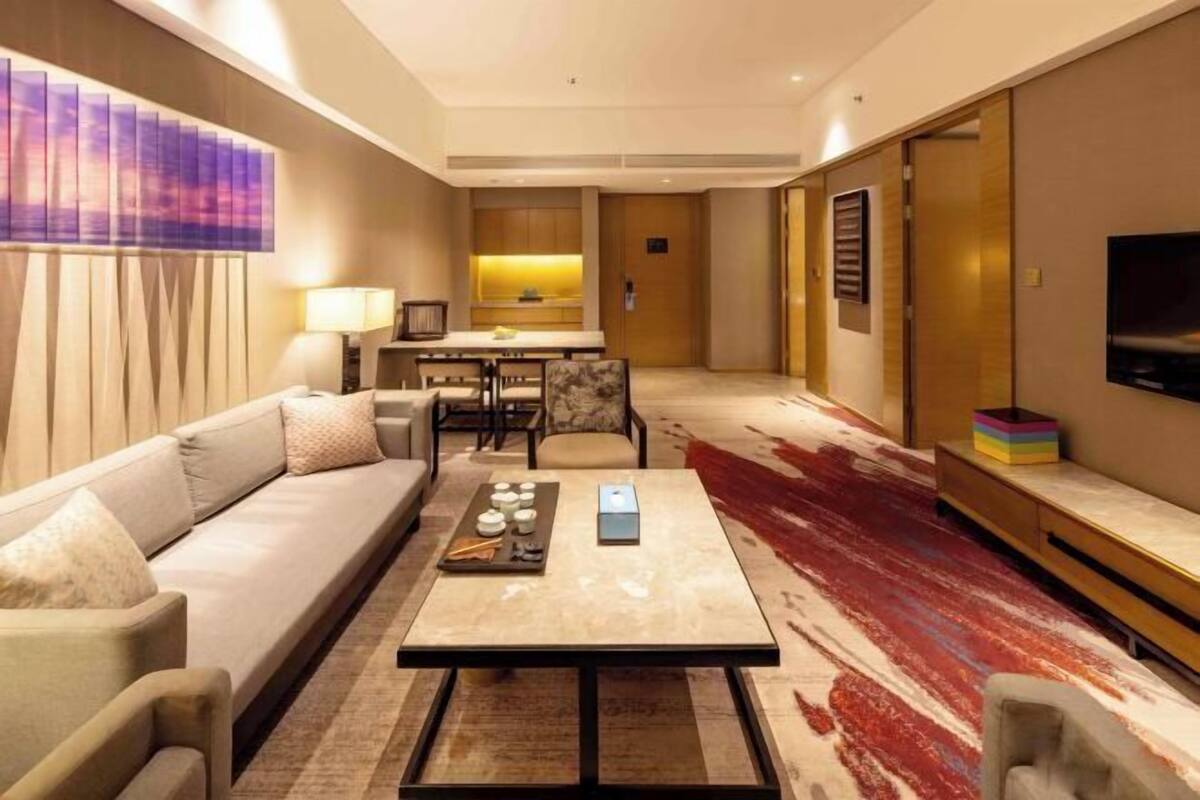 A modern hotel room with a sofa, a dining table, a TV, and a kitchen area.