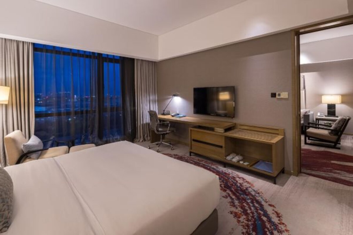 A hotel room with a large bed, a desk with a chair, a TV, and a view of the city at night.