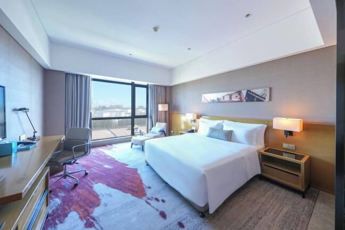 A hotel room with a large bed, a desk, a chair, a nightstand, and a window with a city view.