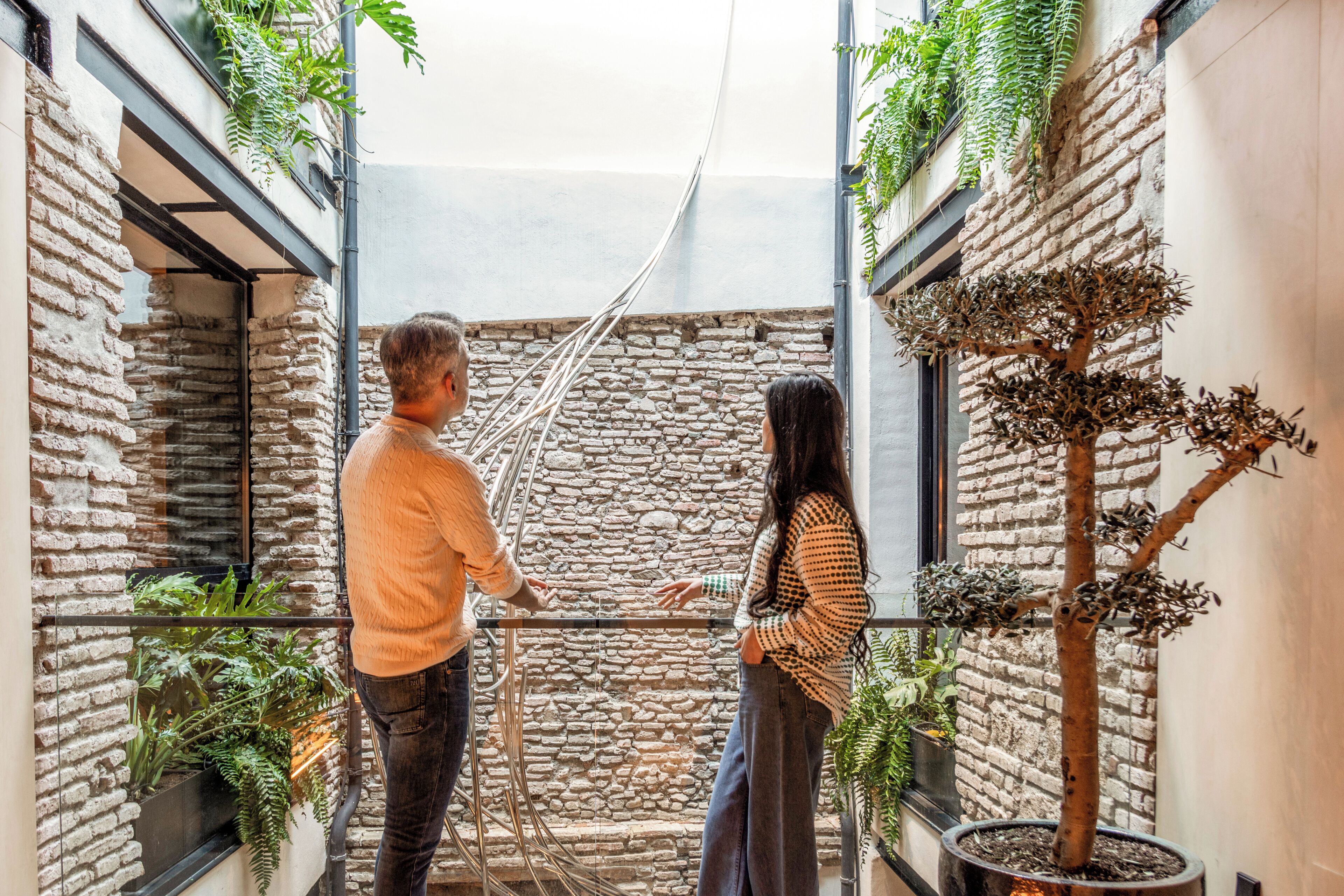 Un espacio interior estrecho con paredes de piedra, barandilla de cristal y plantas en macetas.
